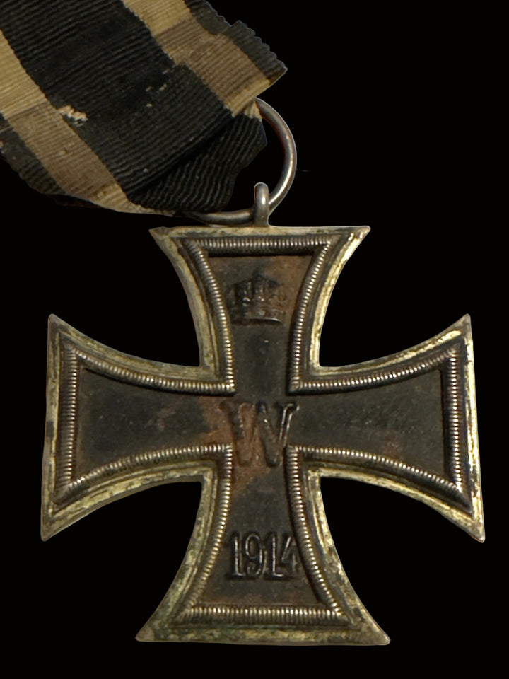 A close-up of a World War I German Iron Cross 1st Class medal with a black and khaki striped ribbon, on a plain off-white background.