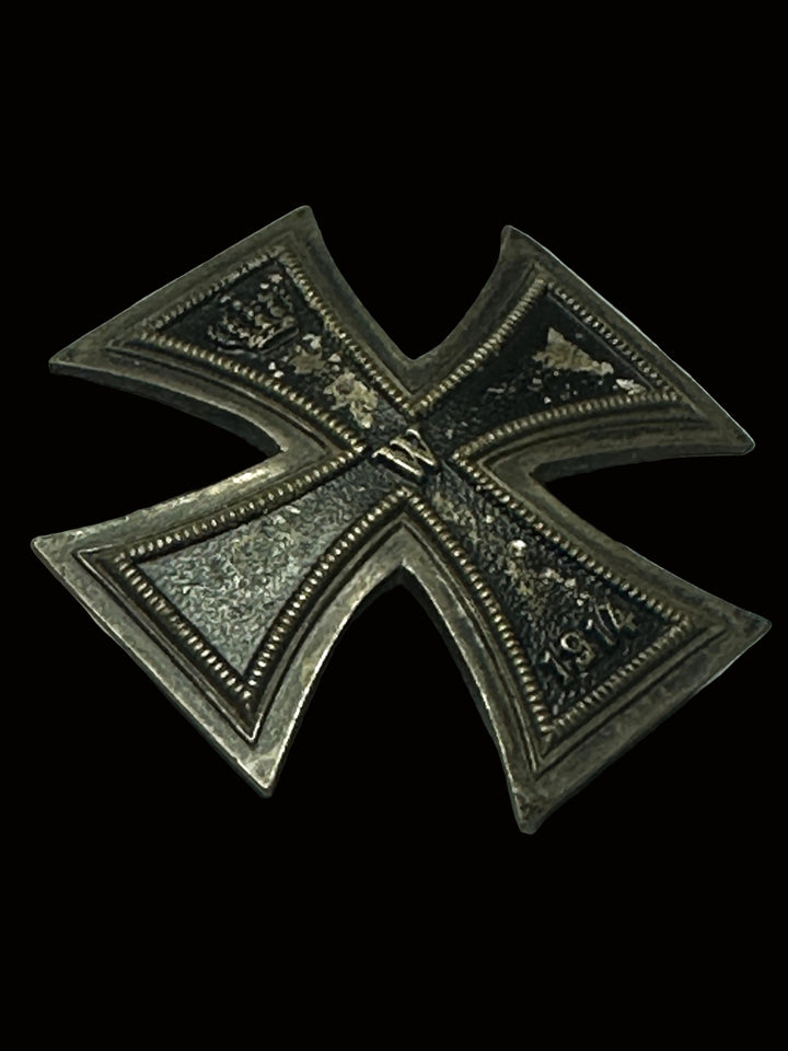 A vintage German Iron Cross 1914 medal pin, featuring a silver-toned metal cross with dark oxidized details and a central crown emblem, resting on a gray textured surface.
