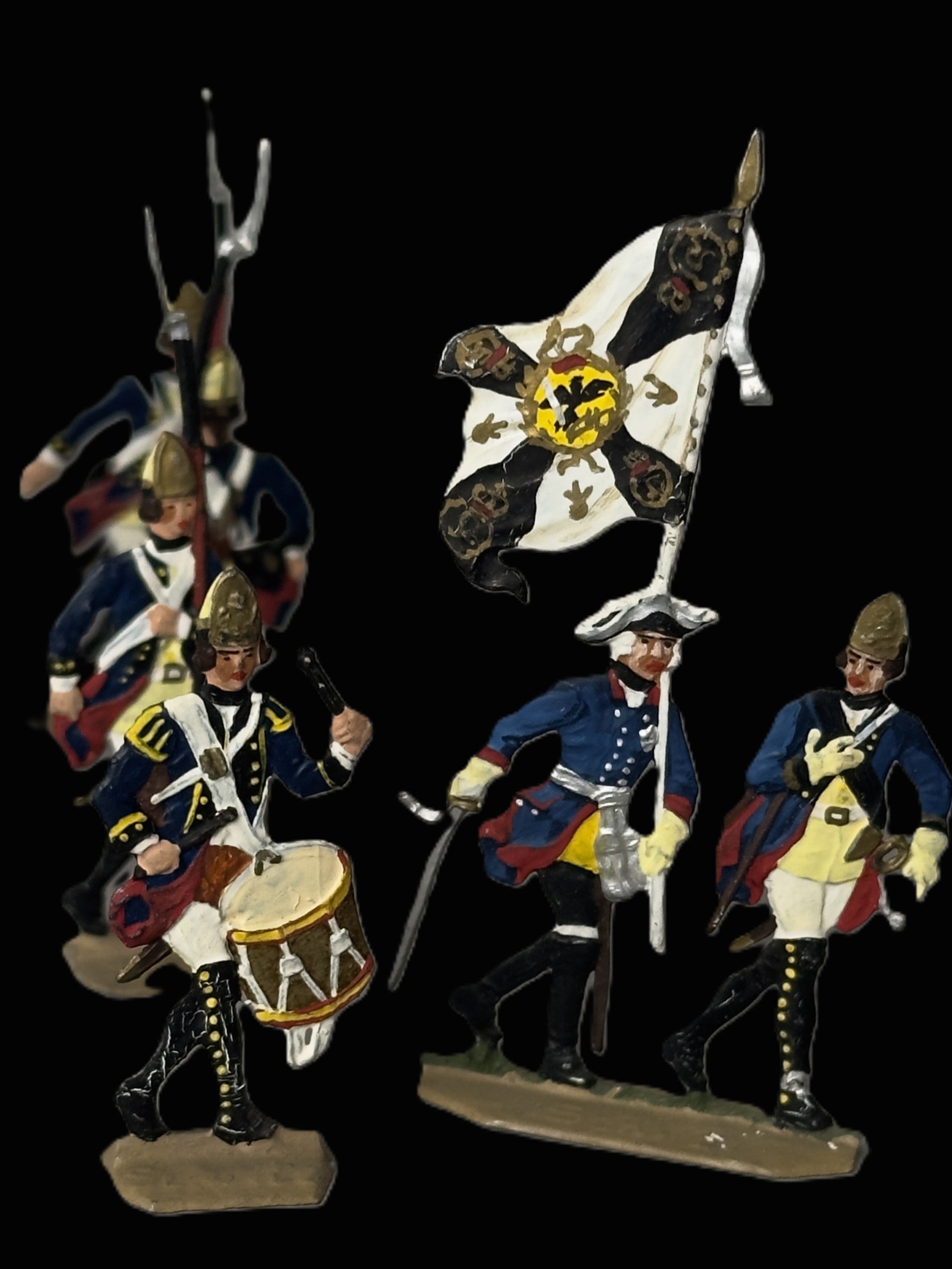 A set of four hand-painted historical military toy figures on a solid black background. The figures include soldiers in blue and white uniforms with yellow accents, a drummer, a flag bearer with a black and white flag featuring a yellow crest, and another soldier carrying a rifle.