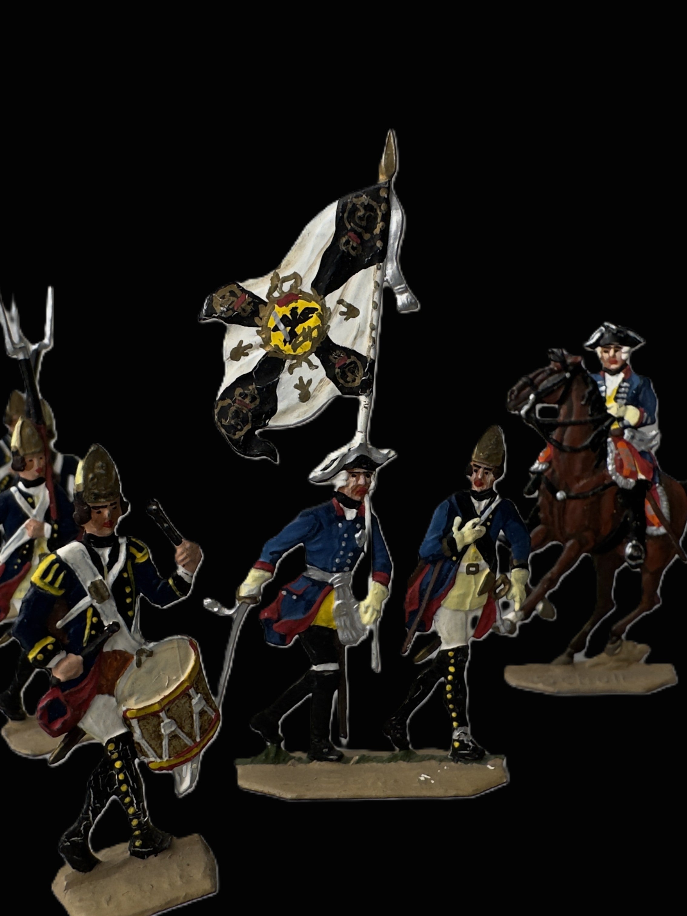 A set of hand-painted historical military toy figures on a solid black background. The figures include soldiers in blue, white, and yellow uniforms, a drummer, a flag bearer, and a mounted soldier on a brown horse.