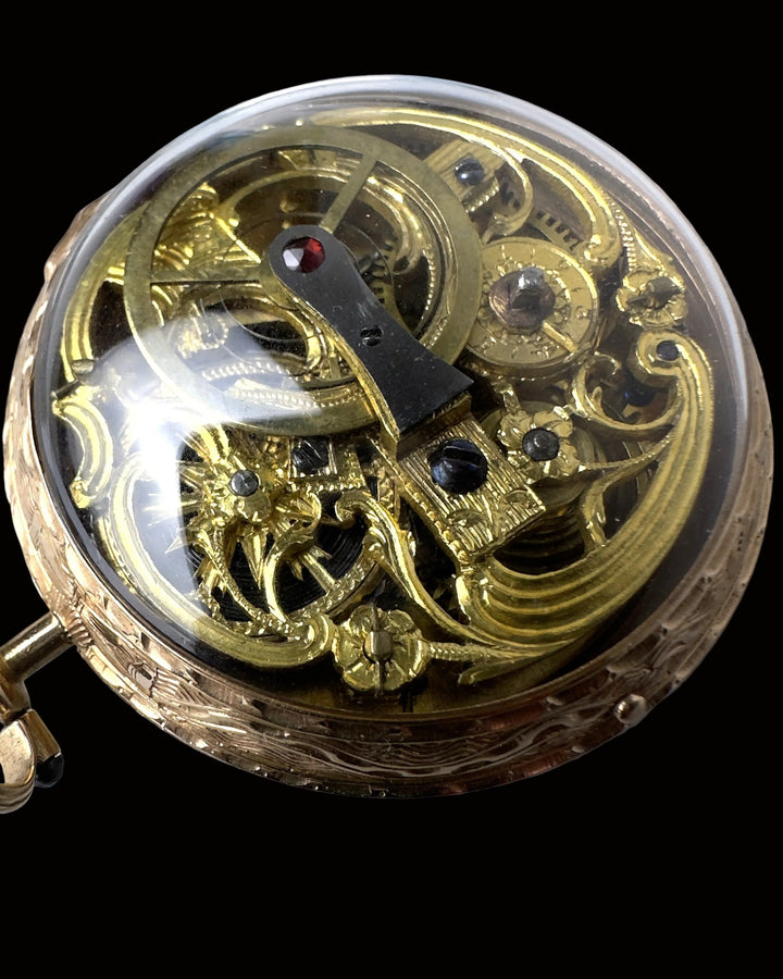 Close-up of an ornate gold-toned mechanical watch movement under a clear protective cover, with visible gears, decorative scrollwork, and a small red jewel.