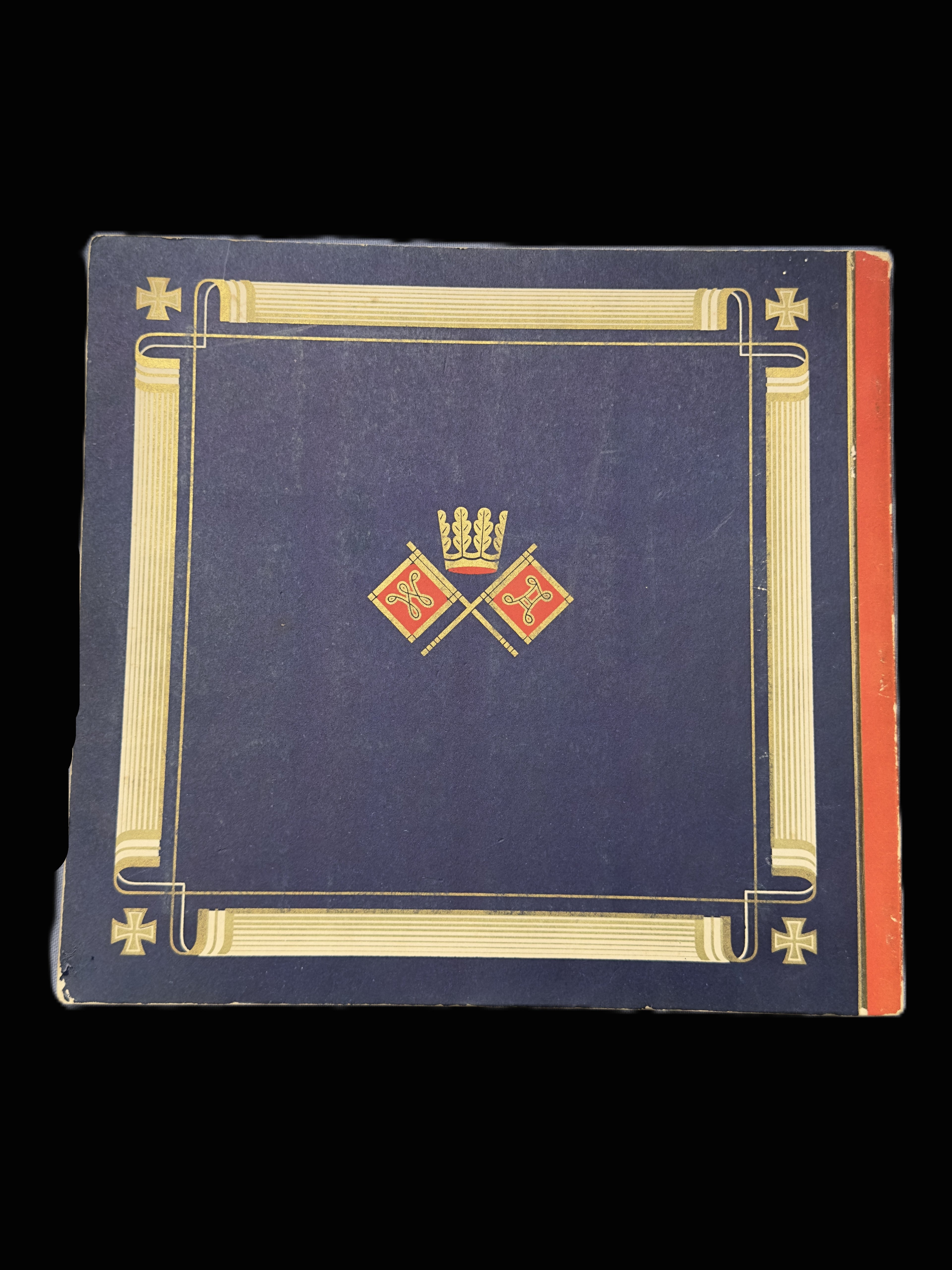 A vintage hardcover book or album with a dark blue cover, featuring a gold decorative border, a central gold and red crest with a crown, and a red-orange spine edge, shown against a solid black background.