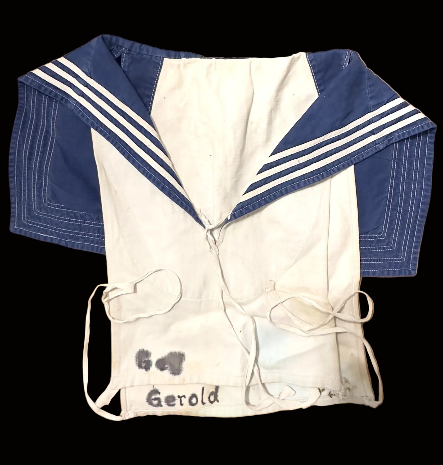 A vintage sailor-style shirt with a white body, blue collar, and white stripes, laid flat on a tan cardboard background. The name 'Gerold' is handwritten on the front.