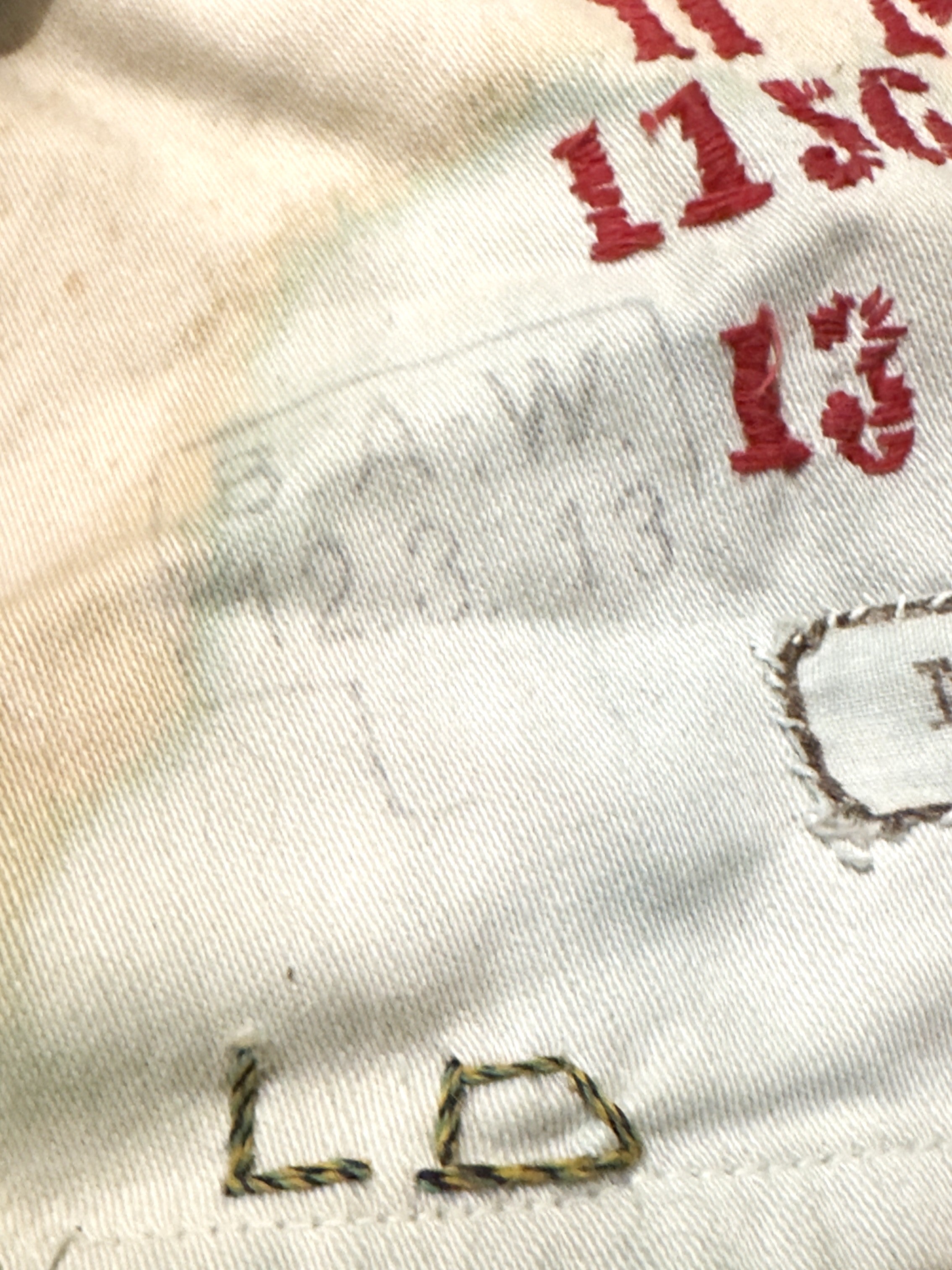 Close-up of a white or off-white fabric surface with red embroidered numbers, a faint gray stamped or printed mark, and partial multicolor stitched lettering.
