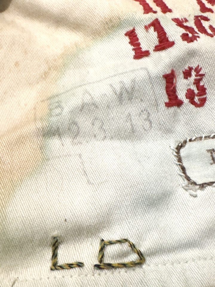 Close-up of a white or off-white fabric surface with red embroidered numbers, a faint gray stamped or printed mark, and partial multicolor stitched lettering.