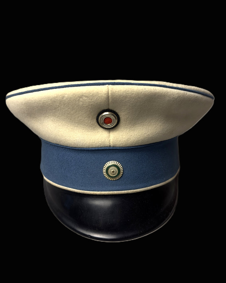A vintage military peaked cap with a cream-colored crown, a blue fabric band, a black leather visor, and gold-toned metal insignia, shown against a solid black background.