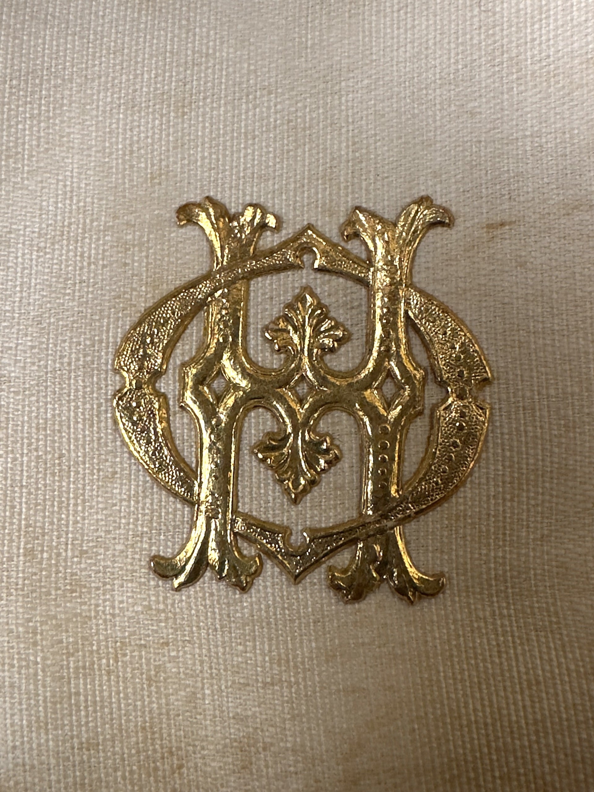 An ornate gold metal monogram emblem featuring an interlocking stylized 'M' and 'H' design, displayed on a textured beige fabric background.