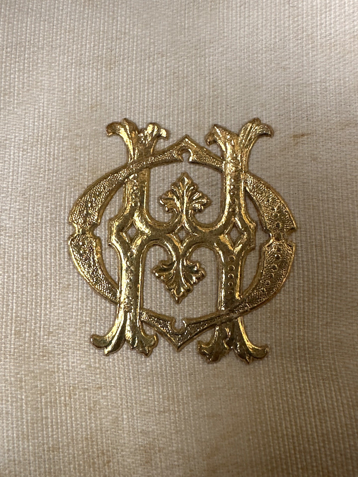 An ornate gold metal monogram emblem featuring an interlocking stylized 'M' and 'H' design, displayed on a textured beige fabric background.