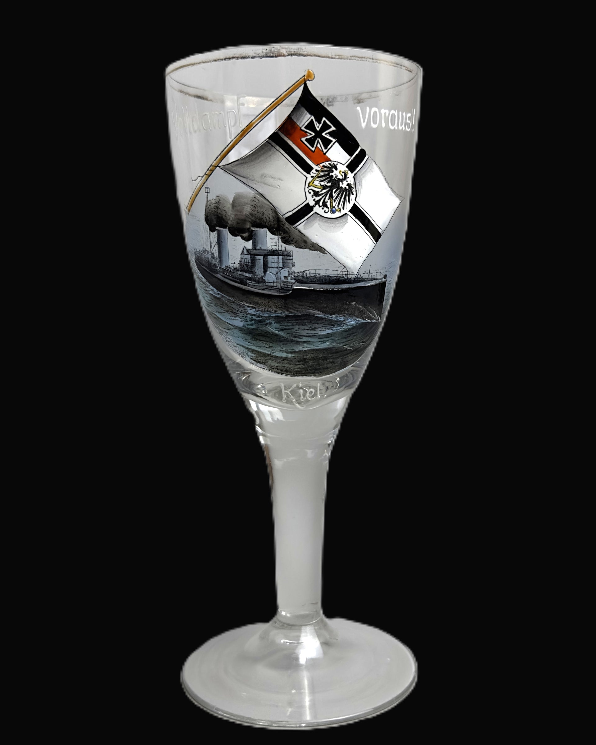 A clear stemmed drinking glass with a printed illustration of a ship on the bowl, featuring a tilted black-and-white flag with a red cross and the word 'VOTCAUS' on a black background.