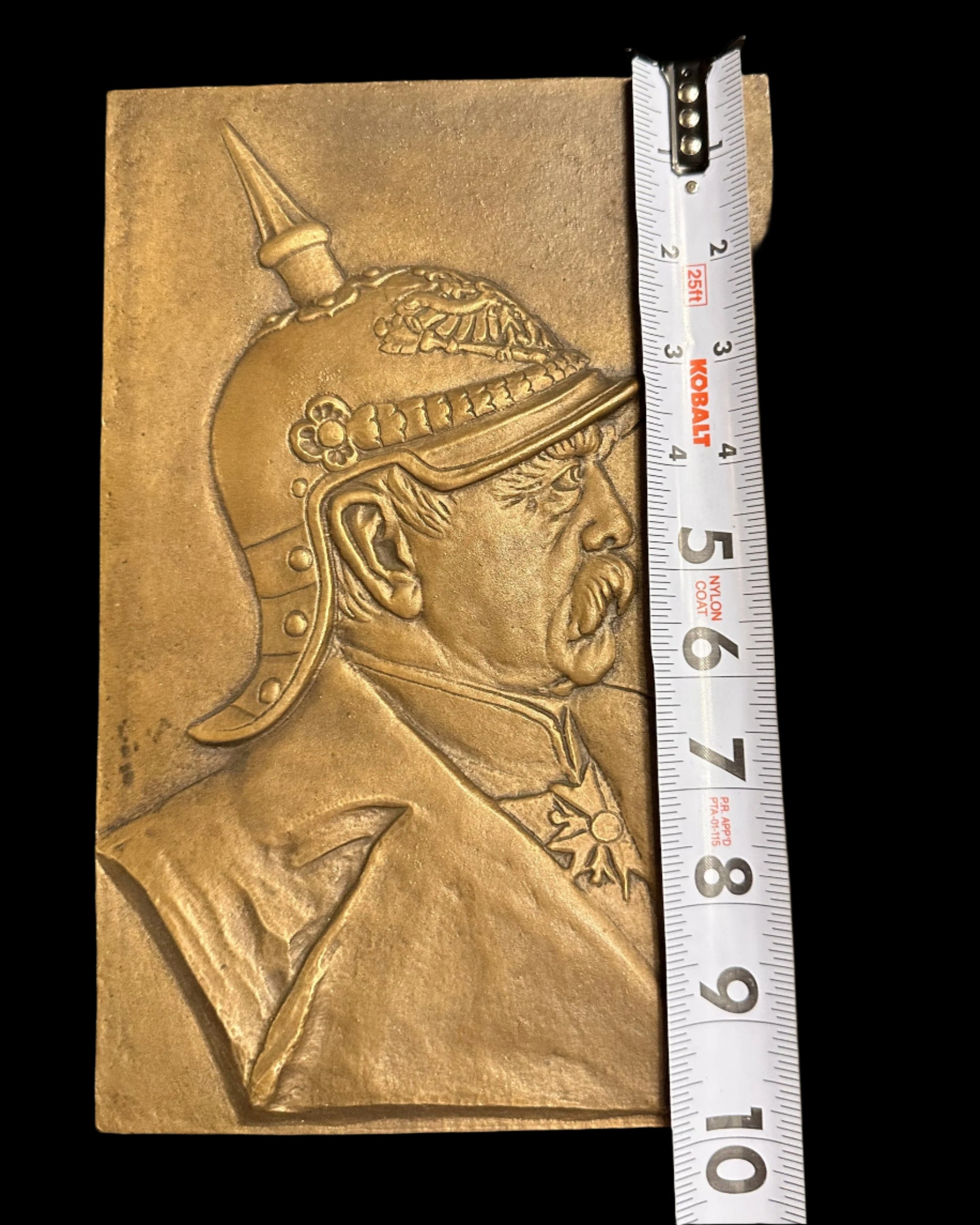 Bronze plaque with embossed design of a helmet, measured against a ruler on a black background.
