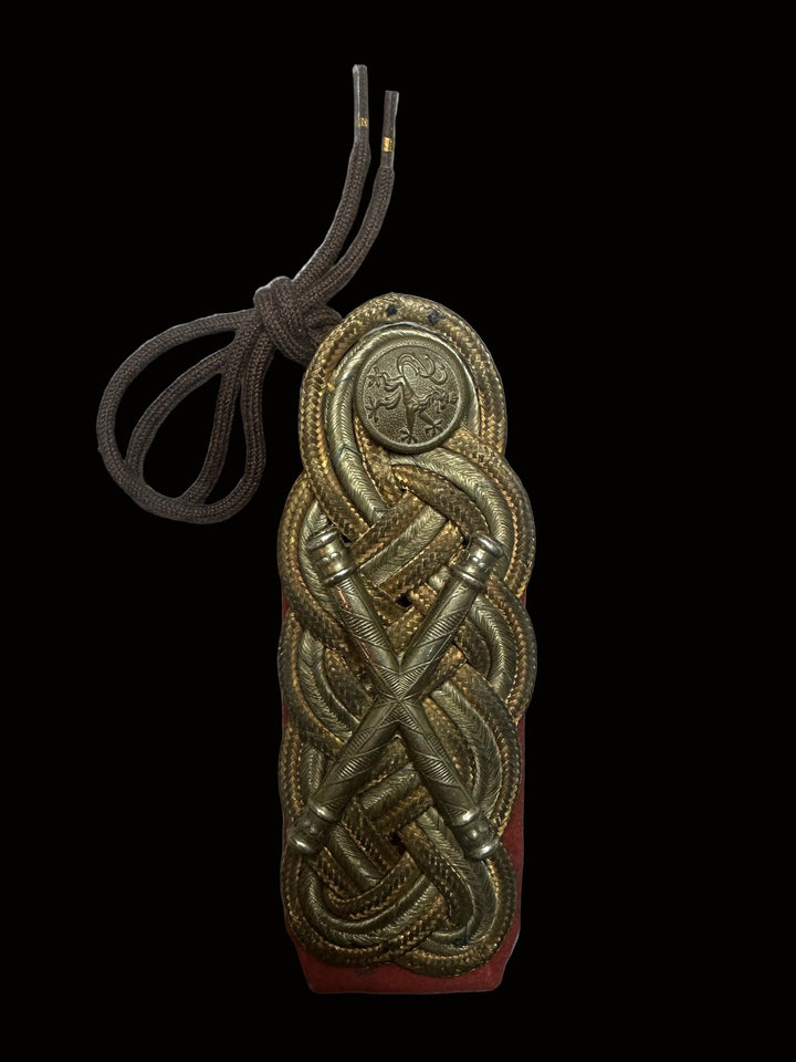 An ornate brass military rank insignia with braided details and a brown cord, displayed on a red backing against a light gray background.