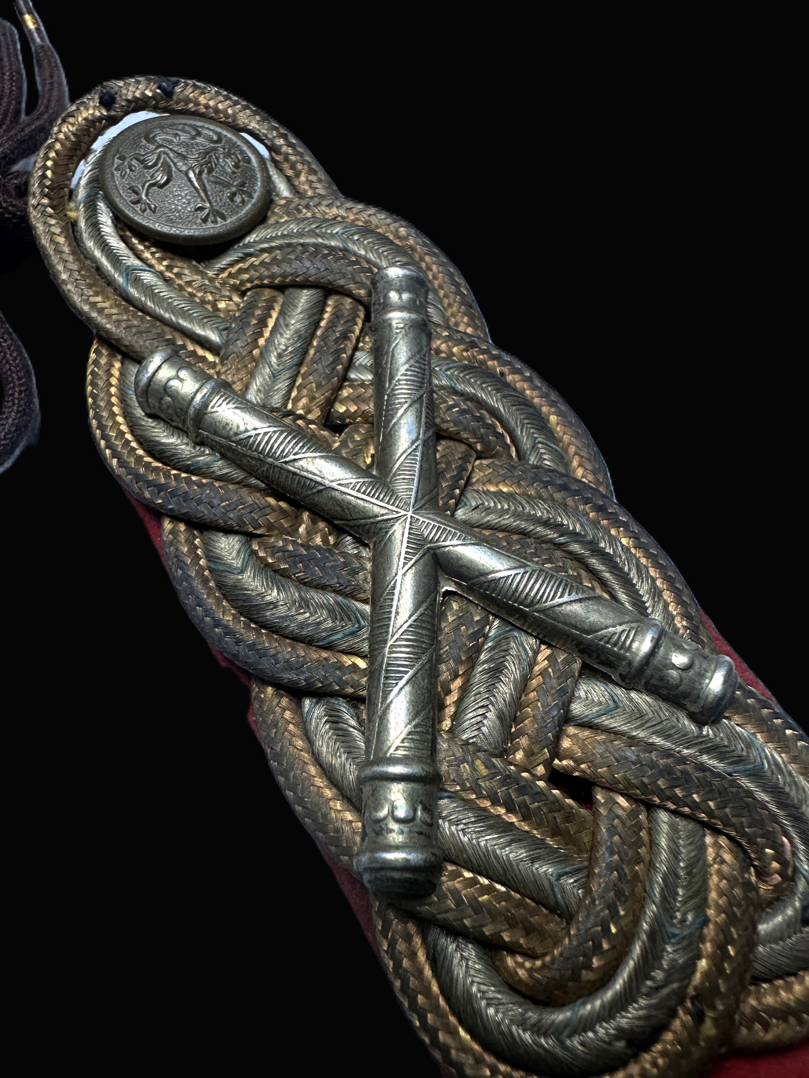 Close-up of an ornate military-style braided cord ornament with metallic silver and gold-toned woven details, a central crossed metal bar, and a round emblem, shown against a black background.