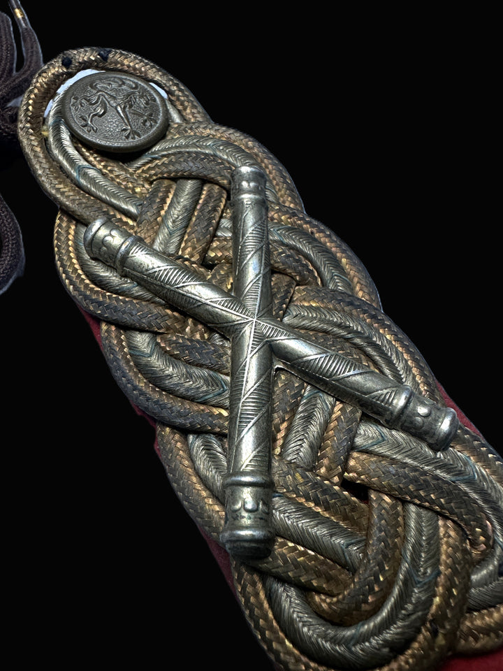 Close-up of an ornate military-style braided cord ornament with metallic silver and gold-toned woven details, a central crossed metal bar, and a round emblem, shown against a black background.