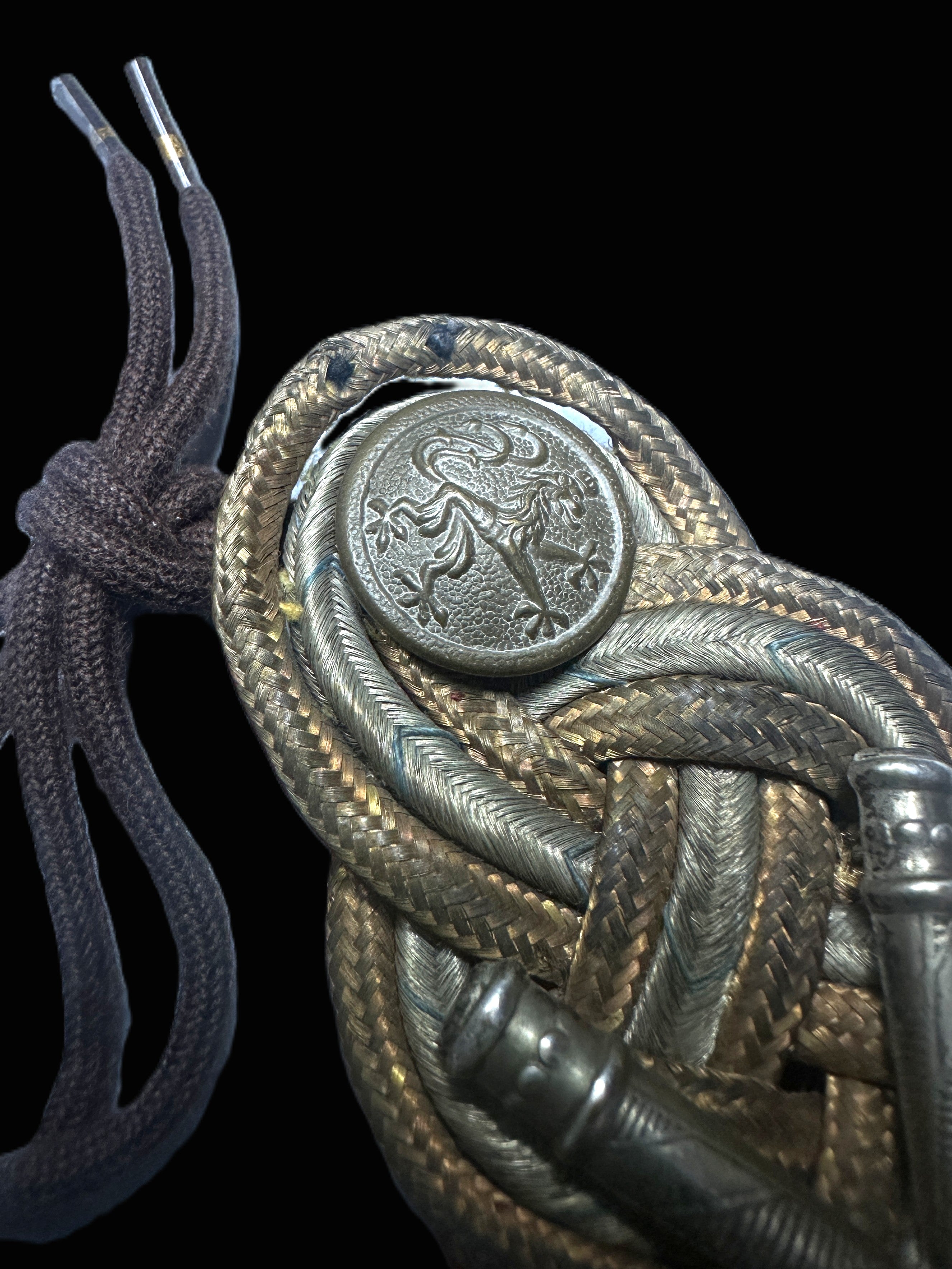 A close-up of an ornate braided leather or rope accessory with a round embossed metal emblem and dark brown cord ties, shown against a solid black background.