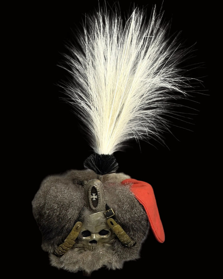 A unique fur hat featuring gray and white fur, a prominent white feather plume, a black feather accent, a red fabric accent, and a skull embellishment on the front.