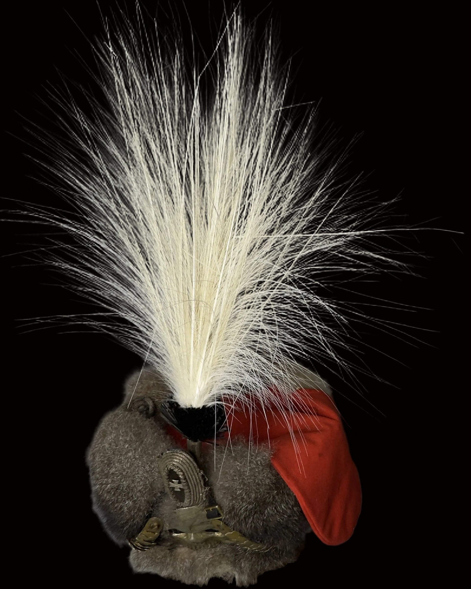A close-up of a fur hat featuring a large white feather plume, gray fur, a red fabric accent, and a gold metal buckle, photographed against a neutral gray background.